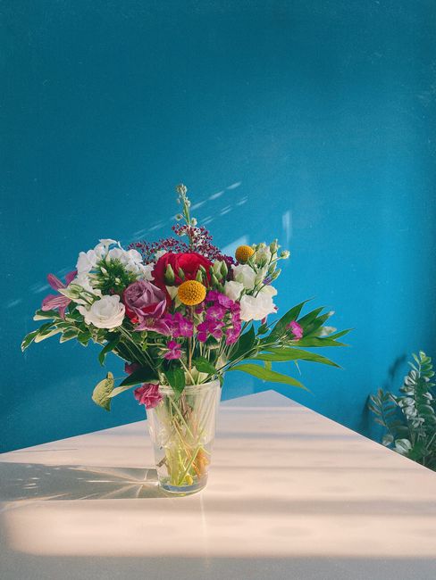 A vibrant floral bouquet arranged in a clear glass vase, featuring a mix of white, pink, purple, and red flowers including roses, lisianthus, and billy balls. The bouquet includes green foliage and is
