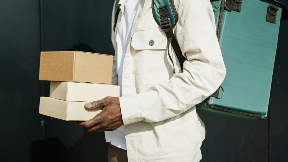 A person dressed in a light-colored jacket holding three rectangular cardboard boxes stacked one on top of the other; the top box is plain brown, the middle is a light cream color, and the bottom box 